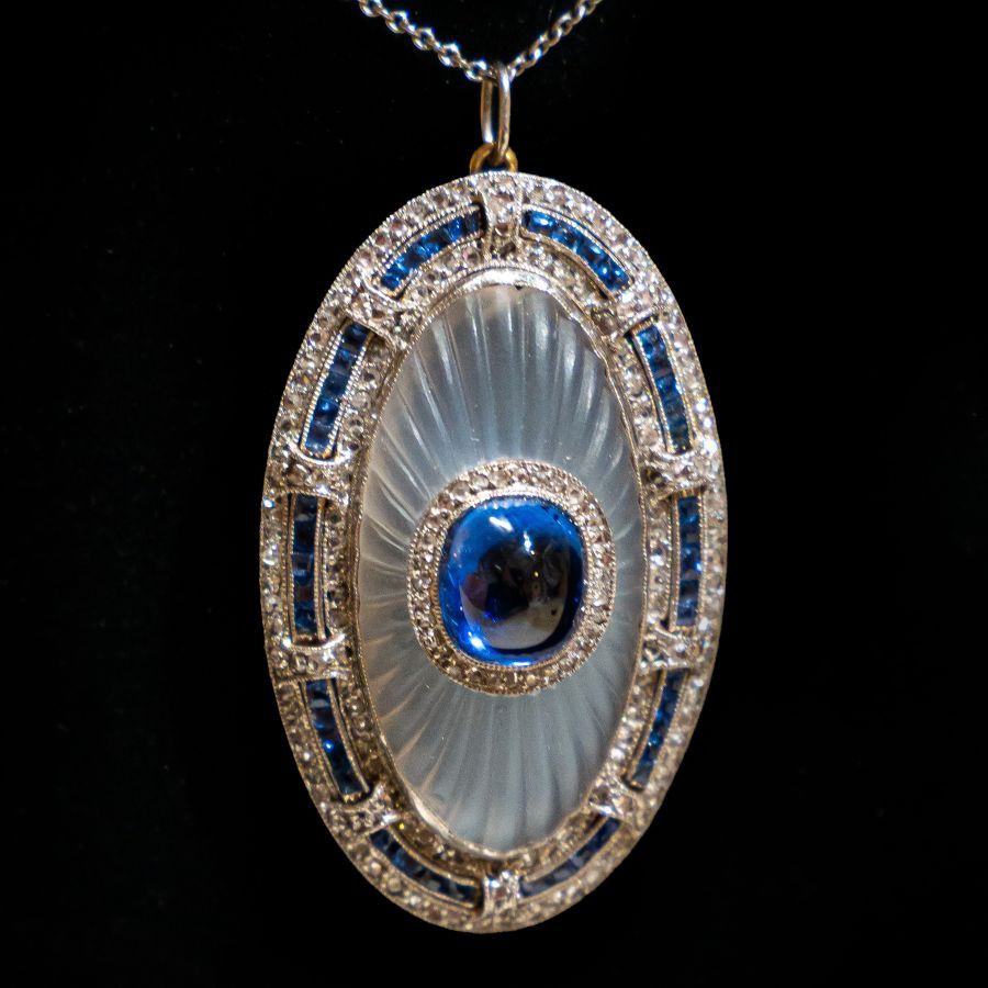 Ein ovaler Anhänger, in der Mitte ein Saphir Cbochon,ca. 5 ct., eingebettet in eine gravierte und mattierte Mondsteinplatte. Der Saphir ist umrahm von kleinen Diamanten und der Mondstein ist umrahmt mit 2 Reihen Diamanten und dazwischen eine Reihe facettierte Saphire. Alles in Platin gefasst.