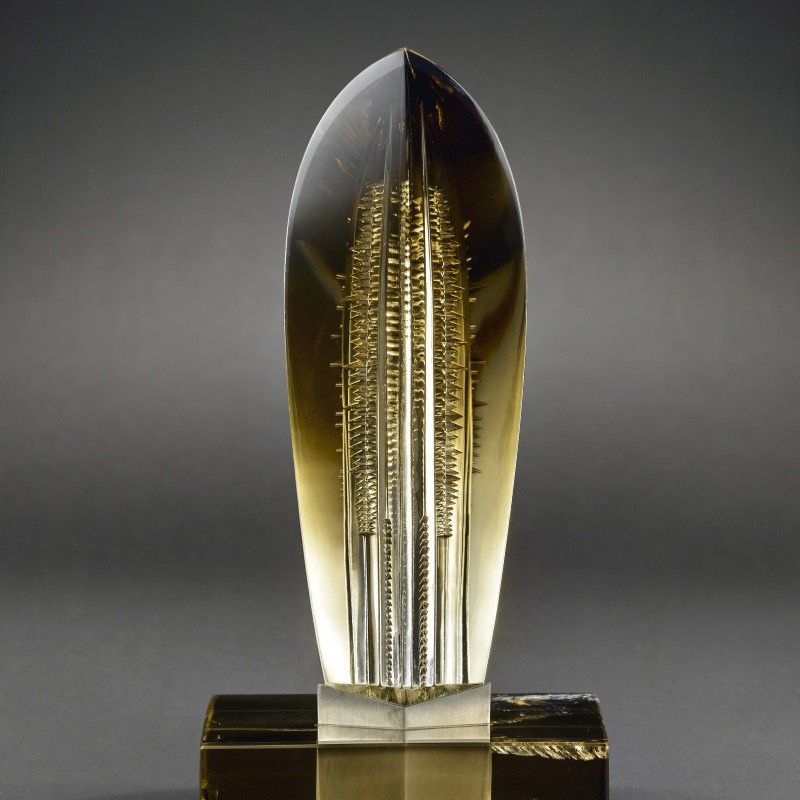 Skulptur aus Citrin mit länglicher, spitz zulaufender Form und inneren, nadelartigen Strukturen auf einem rechteckigen Sockel