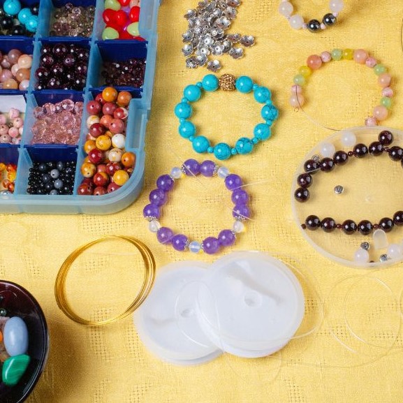 Mehrere bunte Edelsteinarmbänder und eine Aufbewahrungsbox mit verschiedenen Perlen auf gelbem Untergrund  fürs Kinderbasteln