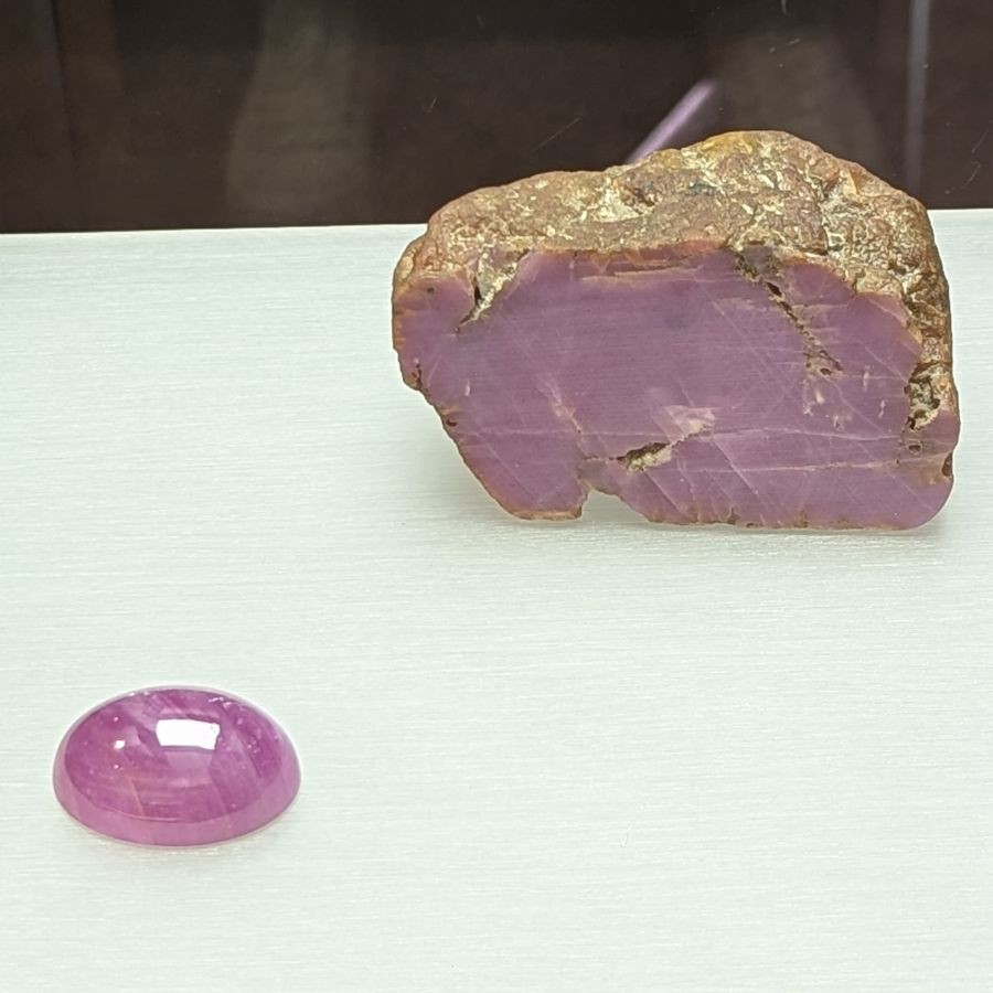 Ein ungeschliffener Rubinrohstein mit rauer Oberfläche und ein polierter Rubin-Cabochon mit glatter, gewölbter Oberfläche liegen nebeneinander auf einer hellen Fläche.